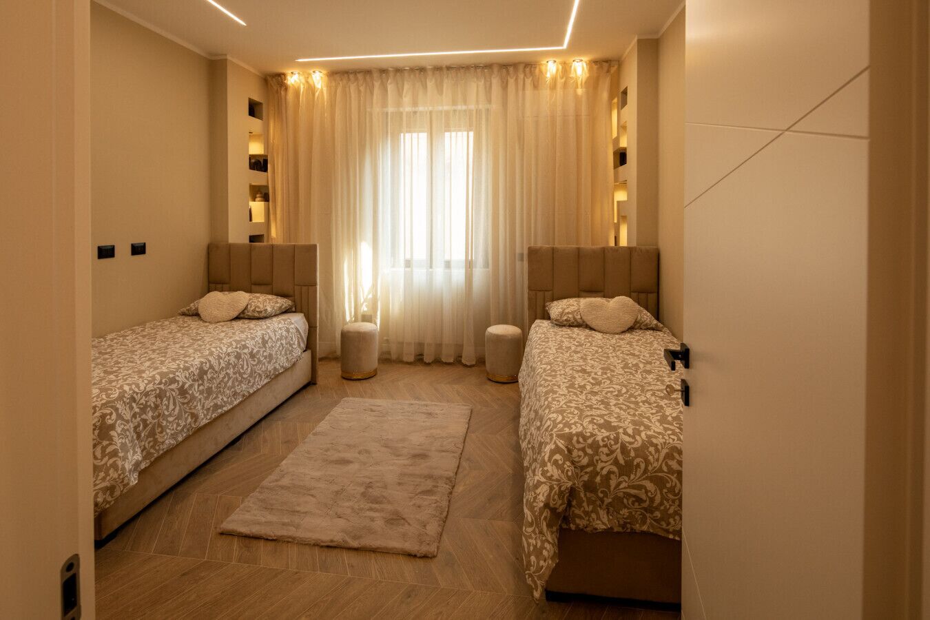 Ein Doppelzimmer mit zwei Betten, einem großen Fenster mit leichtem Vorhang und Holzboden.