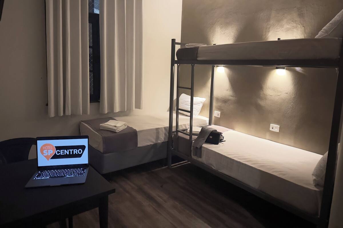 Une chambre avec un lit superposé, un ordinateur portable posé sur un bureau, une fenêtre avec des rideaux et une lampe fixée au mur.