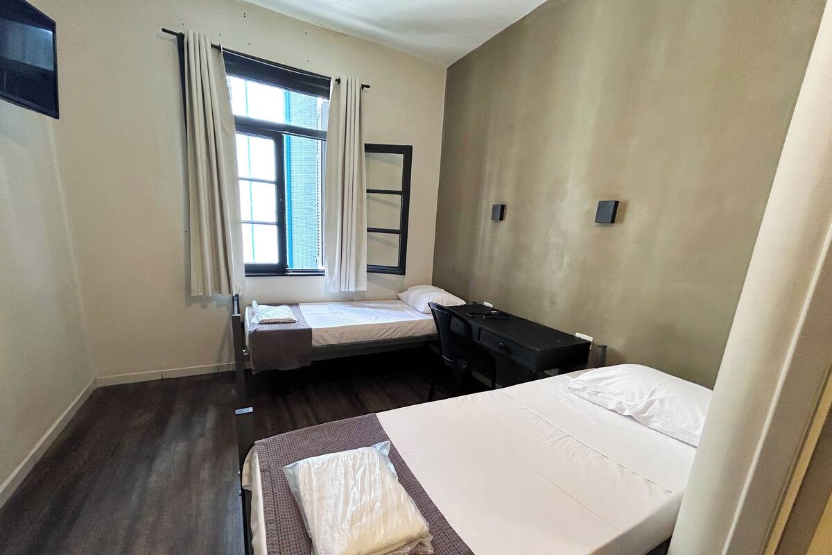 Une petite chambre propre, avec deux lits simples, un bureau, une chaise et une fenêtre avec des rideaux.