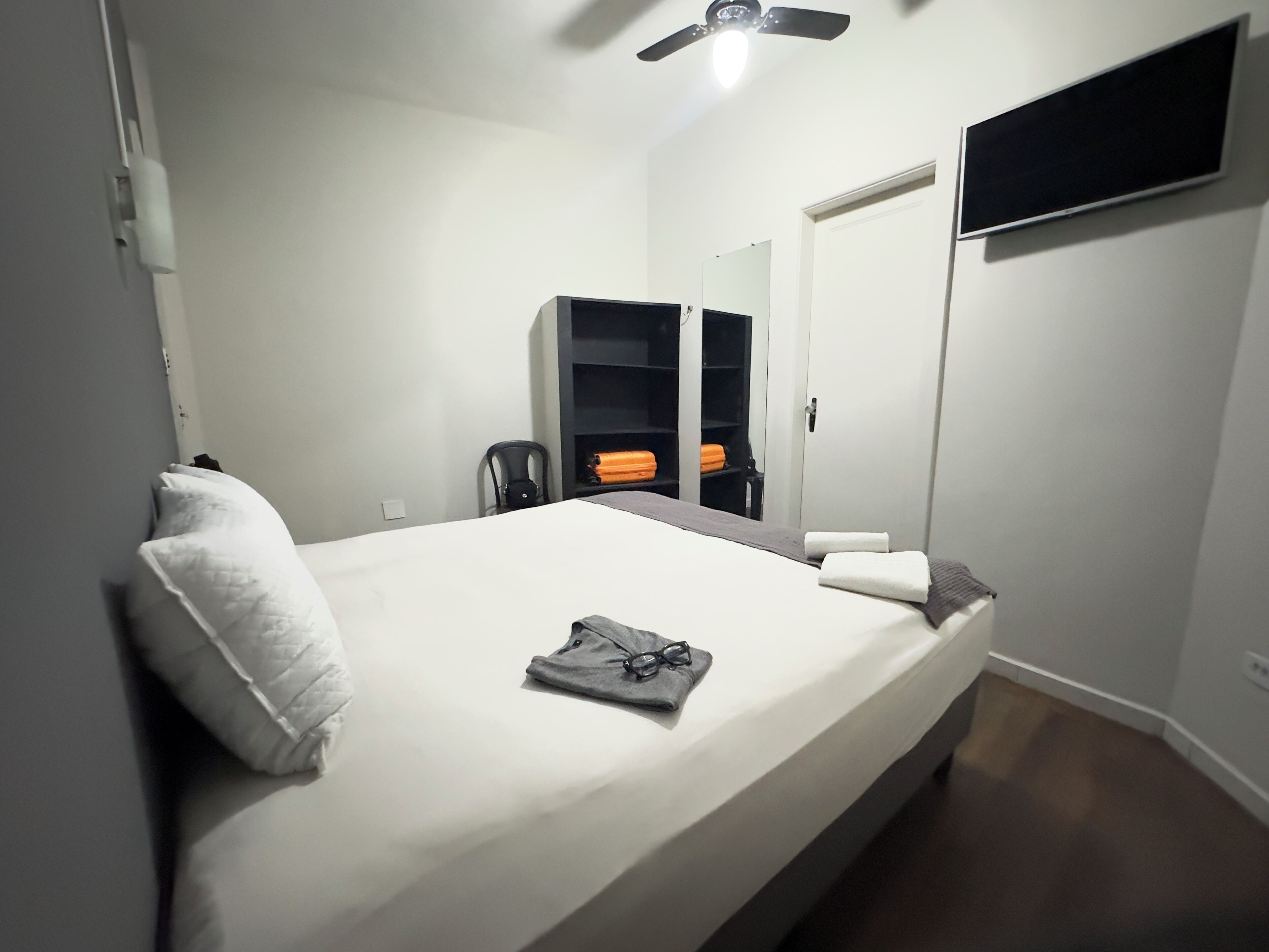 A neatly made bed with a pillow, a folded towel, and a pair of glasses on the bed. There is a chair and a wardrobe in the background.