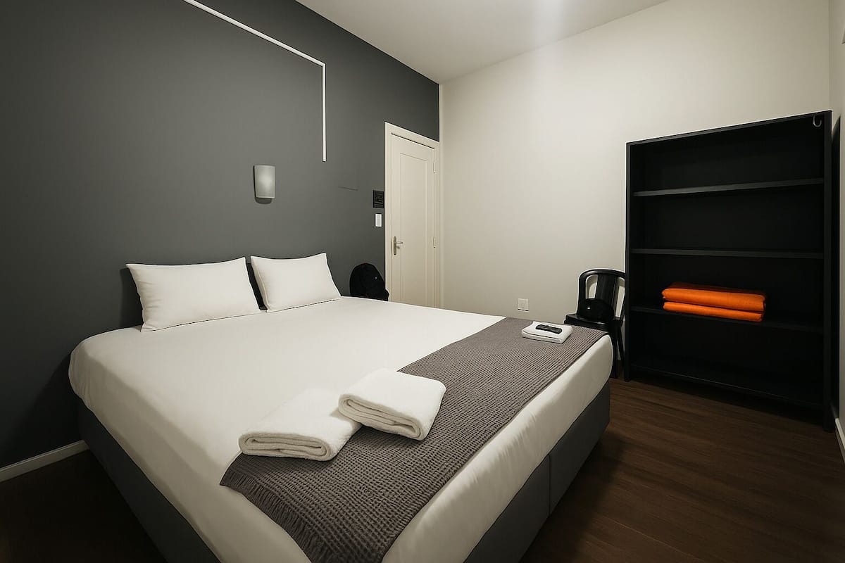 Un lit bien fait, avec du linge de lit blanc, un plaid gris et deux serviettes pliées. Une étagère noire, sur laquelle sont posées des serviettes orange, se trouve à côté du lit.