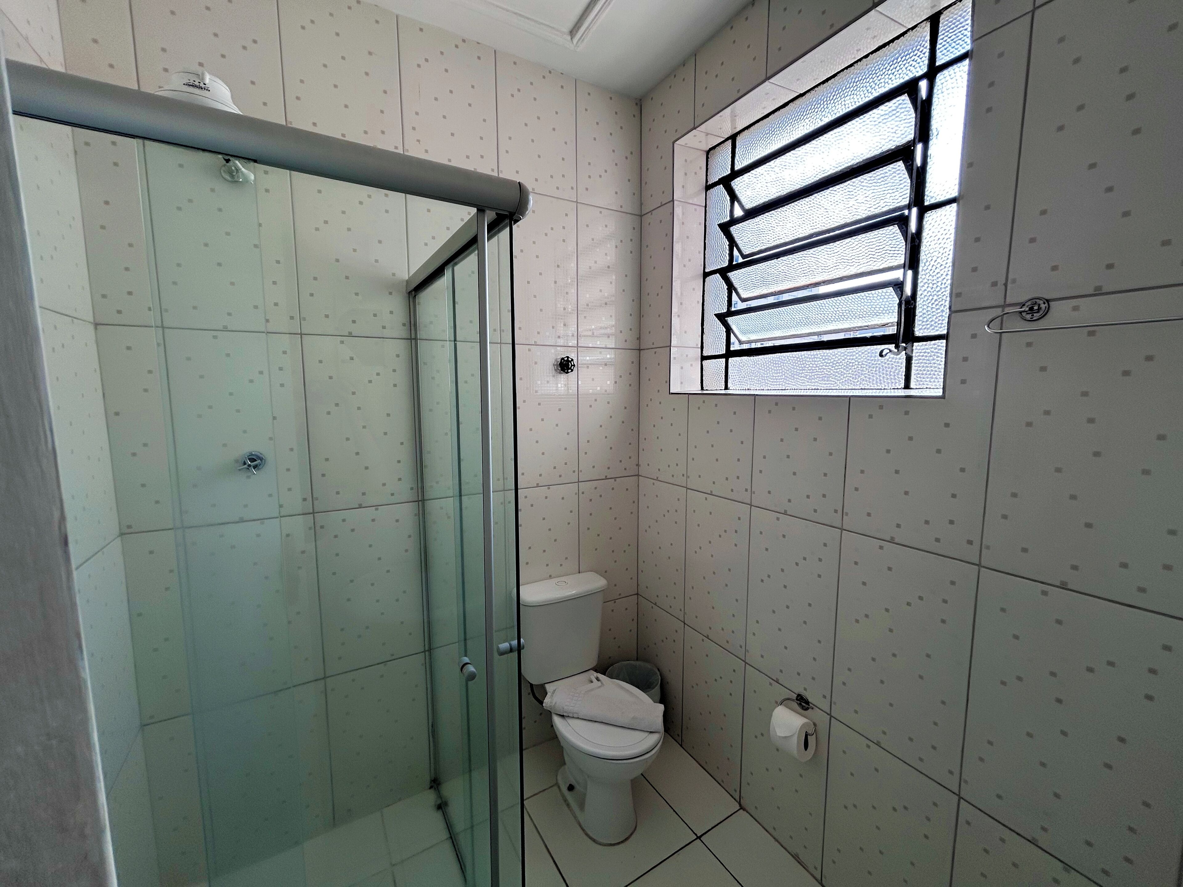 A small, tiled bathroom with a glass shower enclosure, a toilet, and a window with metal bars.
