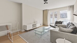A modern living room with a glass coffee table, a grey sofa, a white TV stand, and a dining area with a white table and metal chairs. - Functional Belgravia 2BR Flat Near Victoria (London)