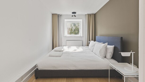 A modern bedroom with a bed, a bedside table, and a window with curtains. - Functional Belgravia 2BR Flat Near Victoria (London)