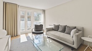 A modern living room with a sofa, a glass coffee table, a leather chair, and a large window with curtains. - Functional Belgravia 2BR Flat Near Victoria (London)