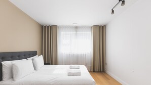 A modern hotel room with a large bed, a wooden headboard, and a view of the city through sheer curtains. - Convenient 2BR/1BA Flat in Belgravia (London)