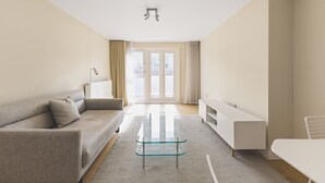 A modern living room with a sofa, a glass coffee table, a white TV stand, and a view through a glass door. - Convenient 2BR/1BA Flat in Belgravia (London)
