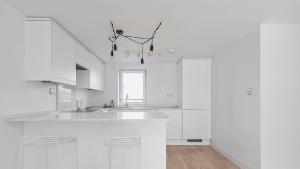 Una cocina moderna con armarios blancos, una isla central con taburetes de bar, una lámpara colgante y un ventanal que proporciona luz natural.