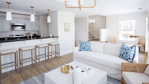 Una cocina moderna con armarios blancos, un microondas y una encimera negra. Hay tres taburetes de bar y un sofá blanco con cojines de estampado azul. Un candelabro cuelga del techo.