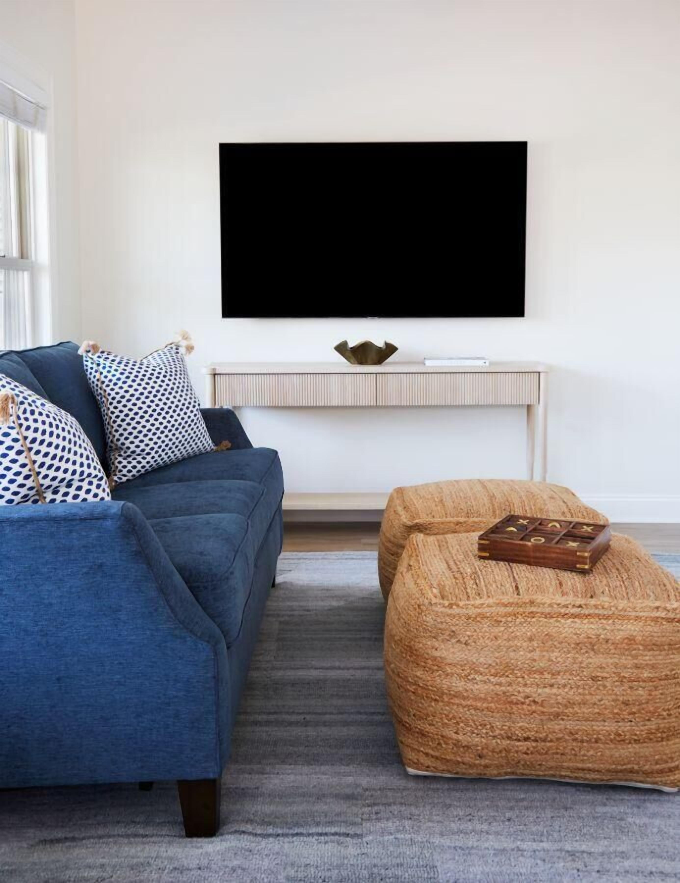 Un salon moderne avec un canapé bleu, des coussins à motifs, un pouf en osier, un téléviseur à écran plat fixé sur une console blanche et un petit bol décoratif.