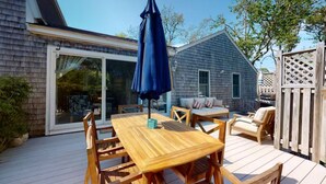 Eine Holzterrasse mit Tisch und Stühlen, einem blauen Sonnenschirm und einer Glastür zu einem überdachten Patio.