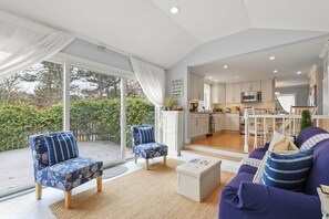 A spacious living area with a large sliding glass door leading to a balcony, a kitchen in the background, and a sofa with striped cushions.