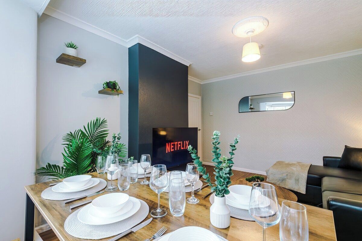 A dining area with a wooden table set for four, a flat-screen TV displaying 'NETFLIX,' and a modern living space with a sofa and a mirror.