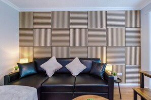 A modern living room with a black leather sofa, two cushions, a small side table with a lamp, and a wooden bench.