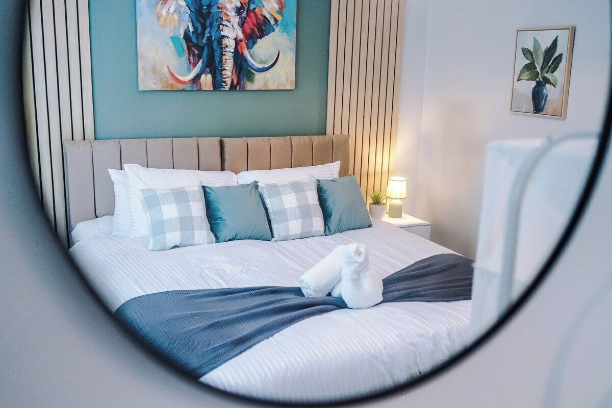 A neatly made bed with a blue blanket and white pillows, a painting of an elephant on the wall, and a framed plant on the side.
