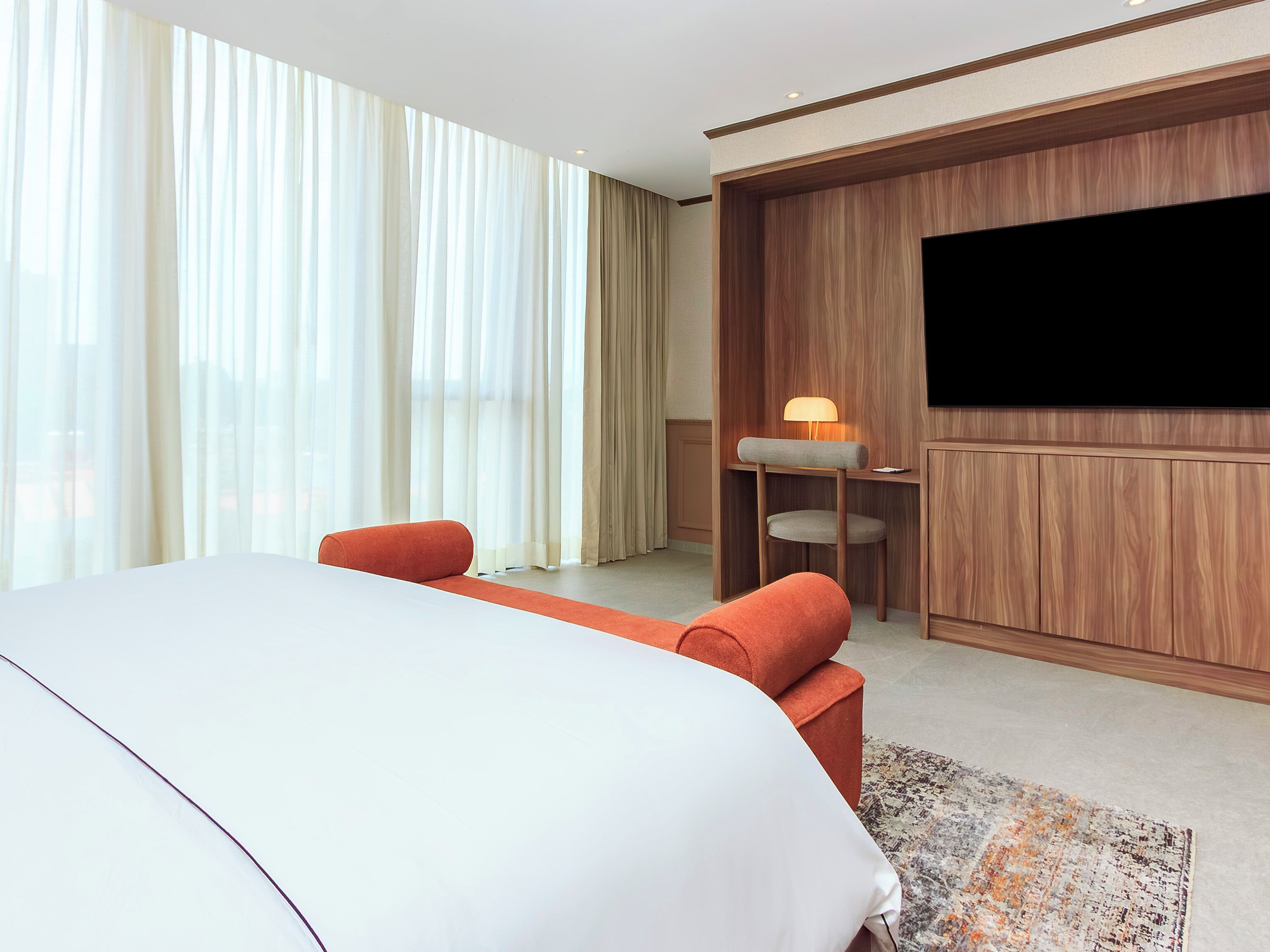 Una habitación de hotel moderna con una cama grande, un escritorio con lámpara, televisor montado en la pared y ventanas amplias con cortinas.