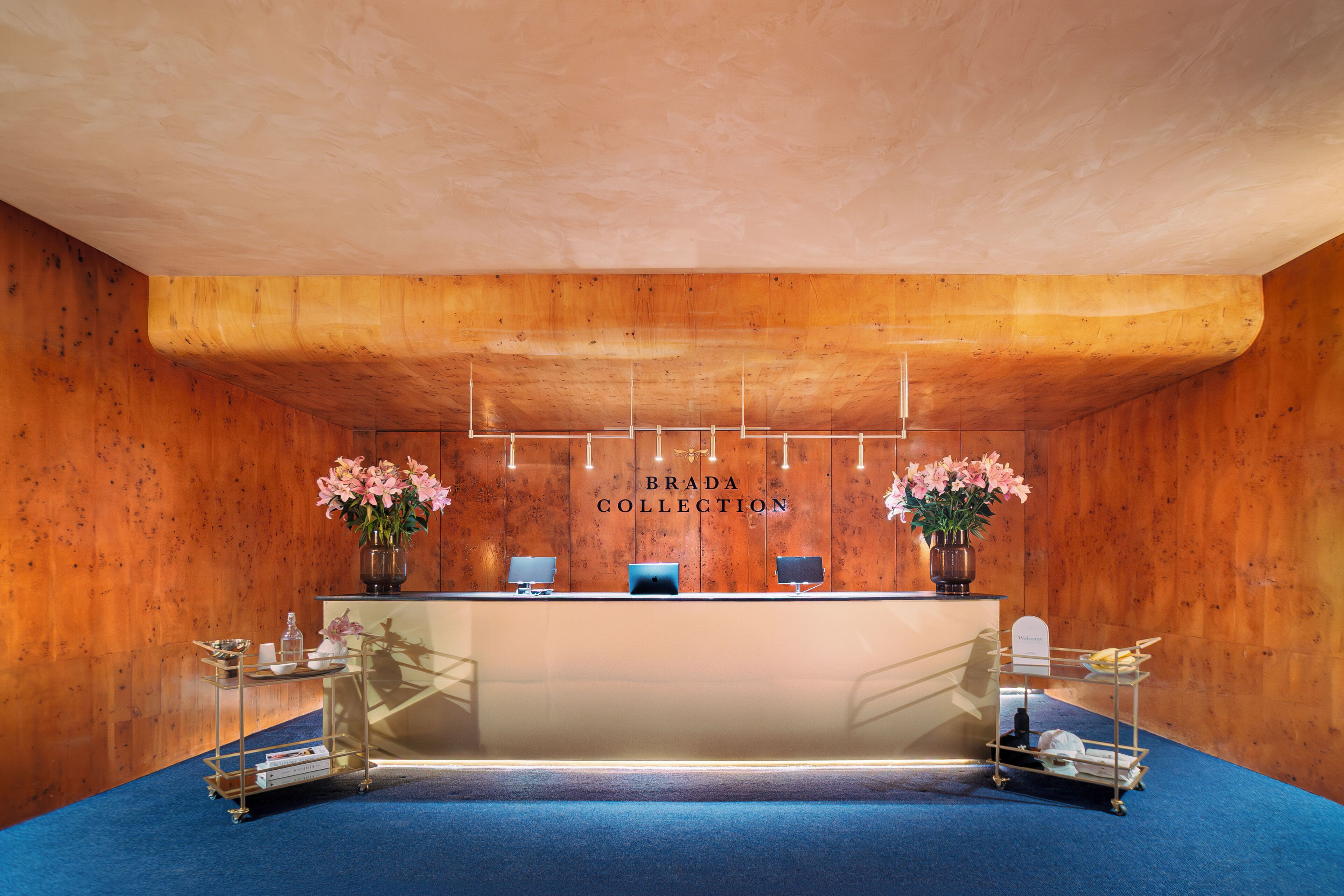A reception desk with a wooden backdrop, two flower arrangements, and a sign reading 'BRADA COLLECTION.'