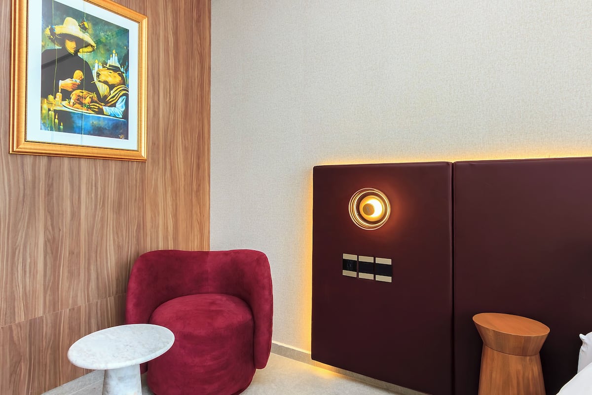 A hotel room with a wooden accent wall, a red armchair, a small round table, a bed with a wooden headboard, and a painting above the headboard.