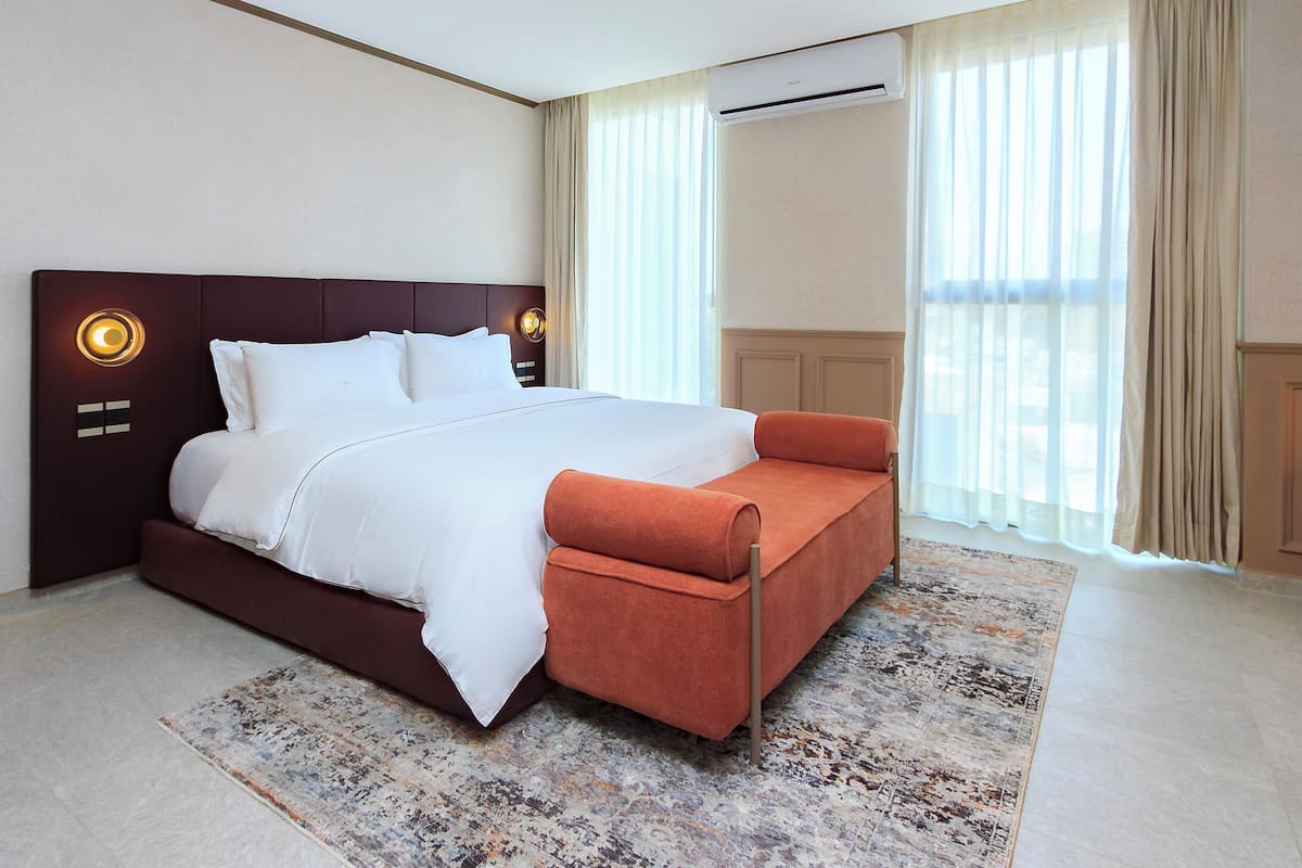 A modern hotel room with a large bed, a sofa, a patterned rug, and a view of the outdoors.