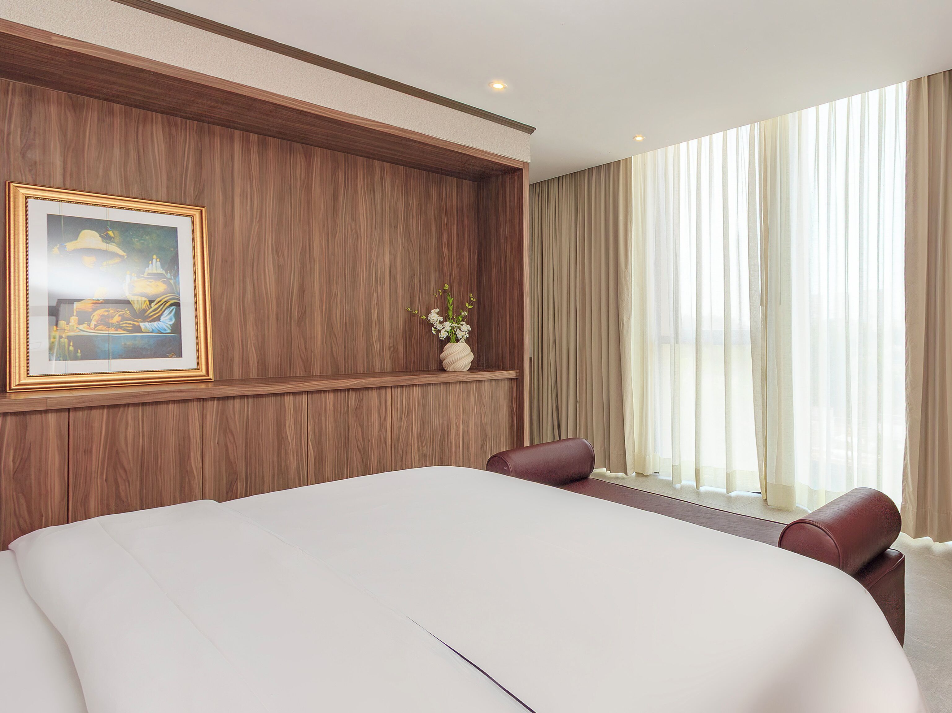 Ein Hotelzimmer mit einem großen Bett, einem gerahmten Bild an der Wand, einer Vase mit Blumen und einem Fenster mit Vorhängen.
