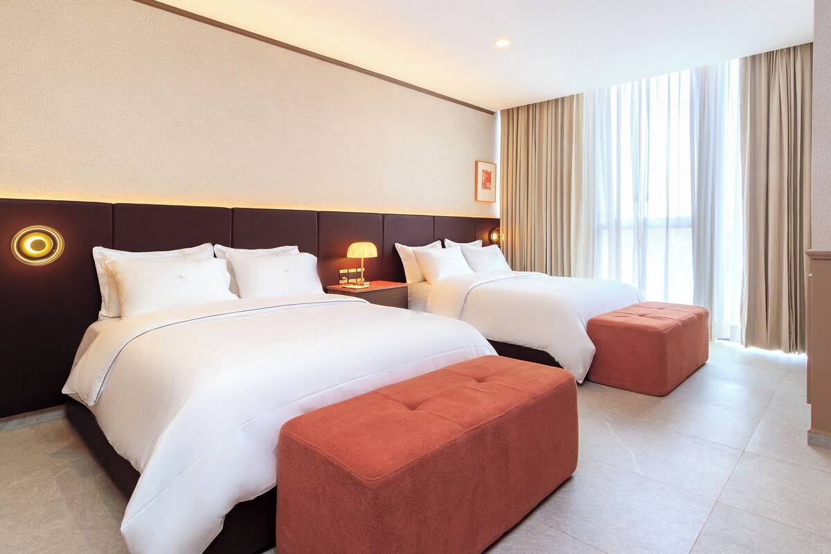 A hotel room with two beds, a nightstand with a lamp, a large window with curtains, and a red ottoman.