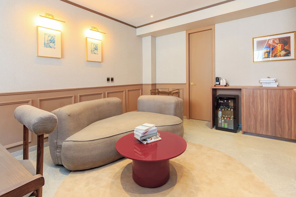 A hotel room with a sofa, a red coffee table, a minibar, and framed artwork on the walls.