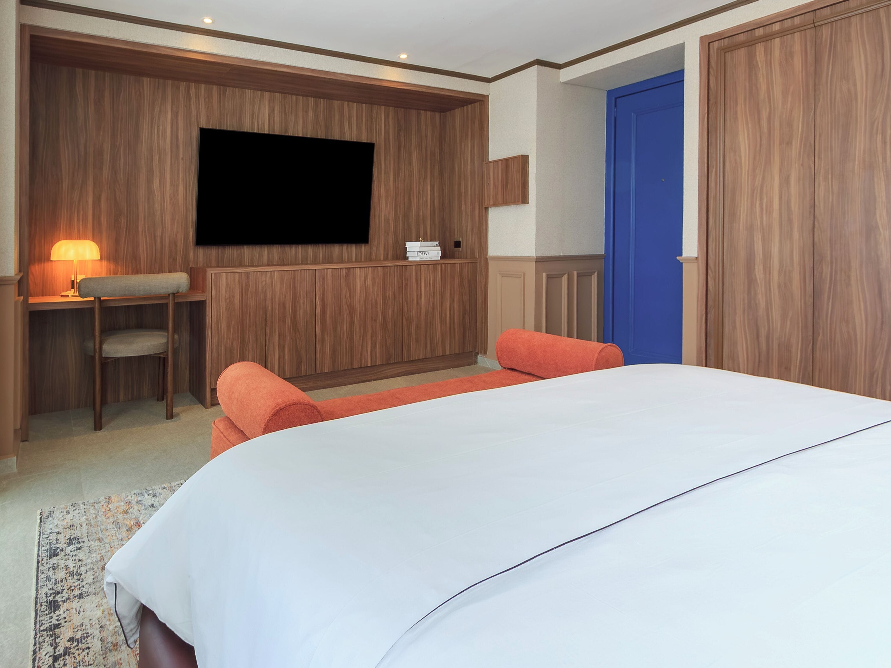 Quarto com cama, escrivaninha com cadeira, televisão na parede e porta azul.