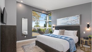 A modern bedroom with a large bed, a view through a window, and a mounted TV. - Seaside 2 bdrm Townhome (Parksville)