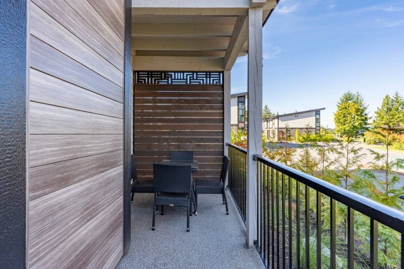 Een balkon met uitzicht op een parkeerplaats en bomen, met een houten wand en een zwarte metalen balustrade.
