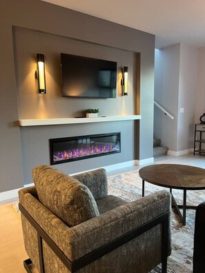 A modern living room with a flat-screen TV mounted above a fireplace, a comfortable sofa, and a round coffee table. - Seaview end unit monthly rates (Parksville)