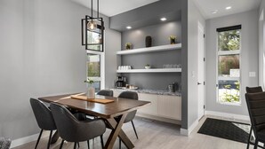 Un comedor moderno con una mesa y sillas de madera, una cocina con armarios y estanterías blancas, y un amplio ventanal que proporciona luz natural.
