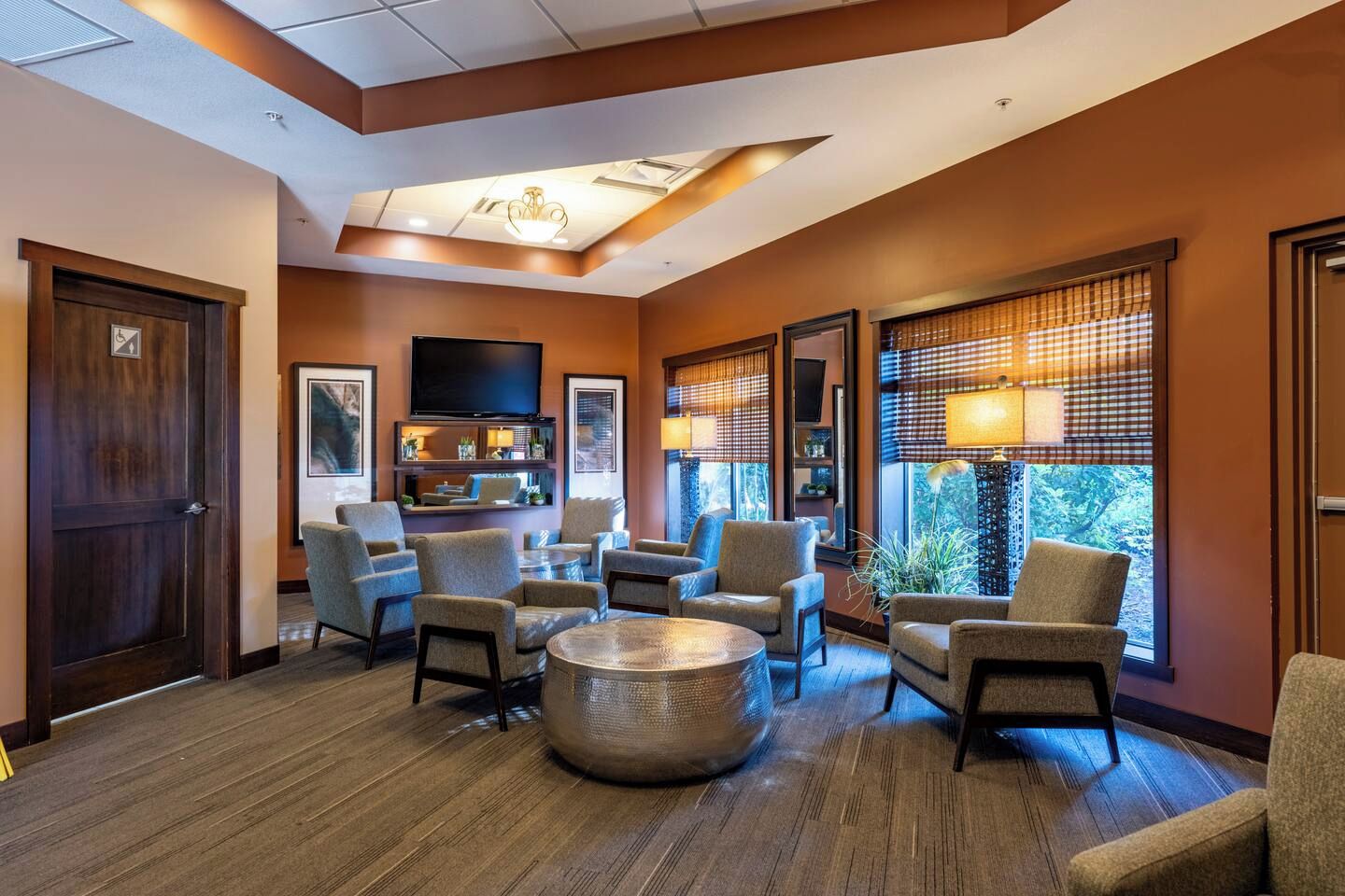 A hotel lobby with a seating area, a television mounted on the wall, and large windows with blinds.