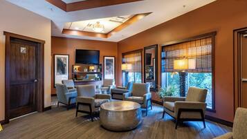 A hotel lobby with a seating area, a television mounted on the wall, and large windows with blinds.