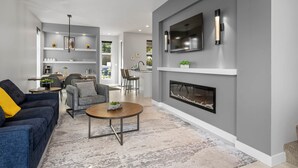 Una sala de estar moderna con chimenea, un televisor montado en la pared y una cocina visible al fondo.