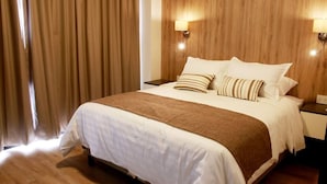 A hotel room with a large bed, wooden headboard, two bedside lamps, and a window with curtains. - Urban Suites (Santa Cruz)