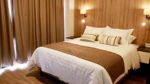 A hotel room with a large bed, wooden headboard, two bedside lamps, and a window with curtains. - Urban Suites (Santa Cruz)