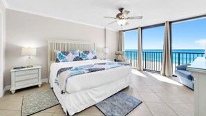 A bedroom with a large bed, a ceiling fan, a nightstand, a lamp, a view of the ocean through large windows, and a balcony with a chair.
