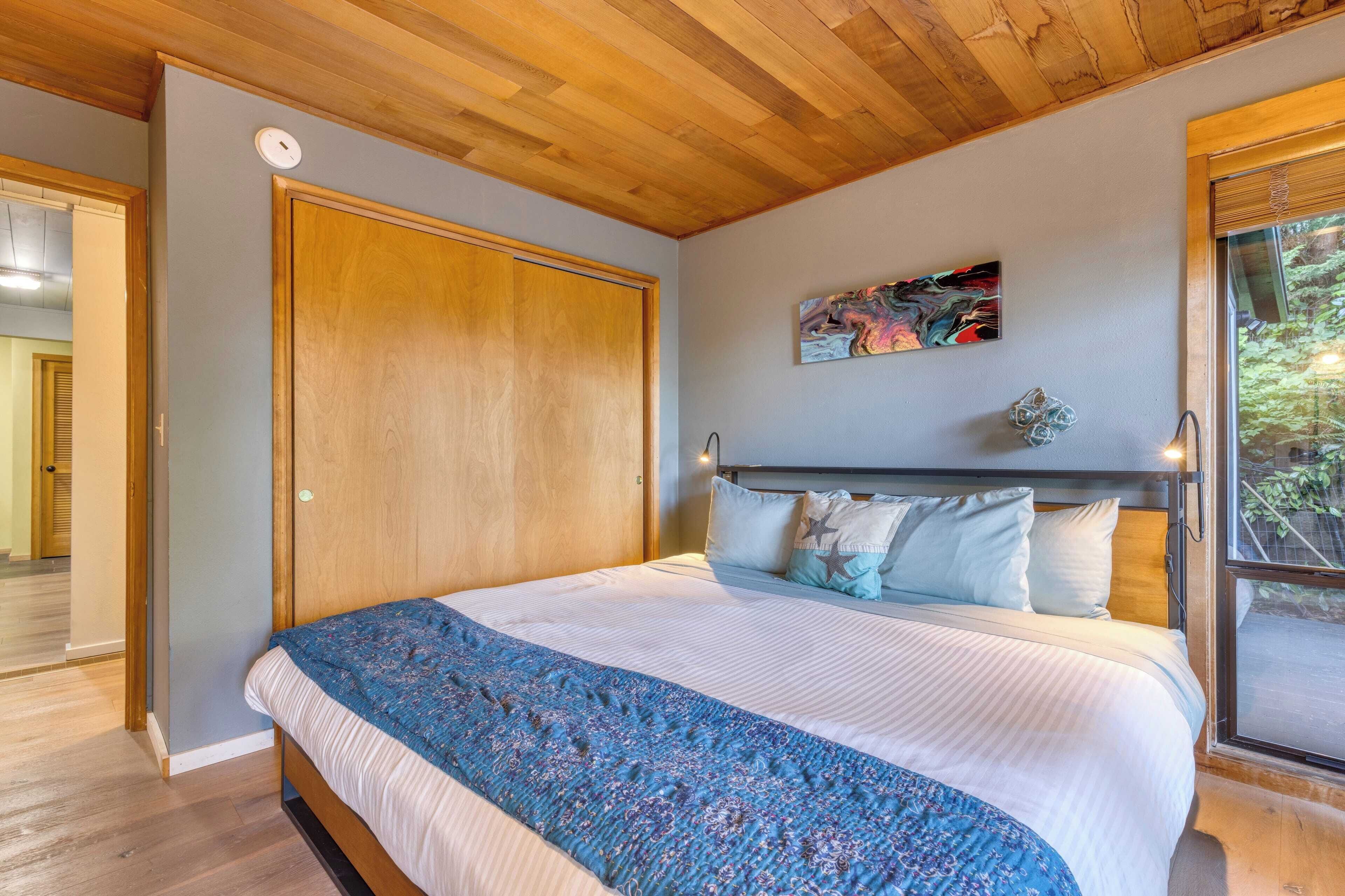 A bedroom with a wooden ceiling, a large bed with a blue patterned blanket, a painting on the wall, and a window with a view of greenery.