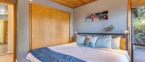 A bedroom with a wooden ceiling, a large bed with a blue patterned blanket, a painting on the wall, and a window with a view of greenery.