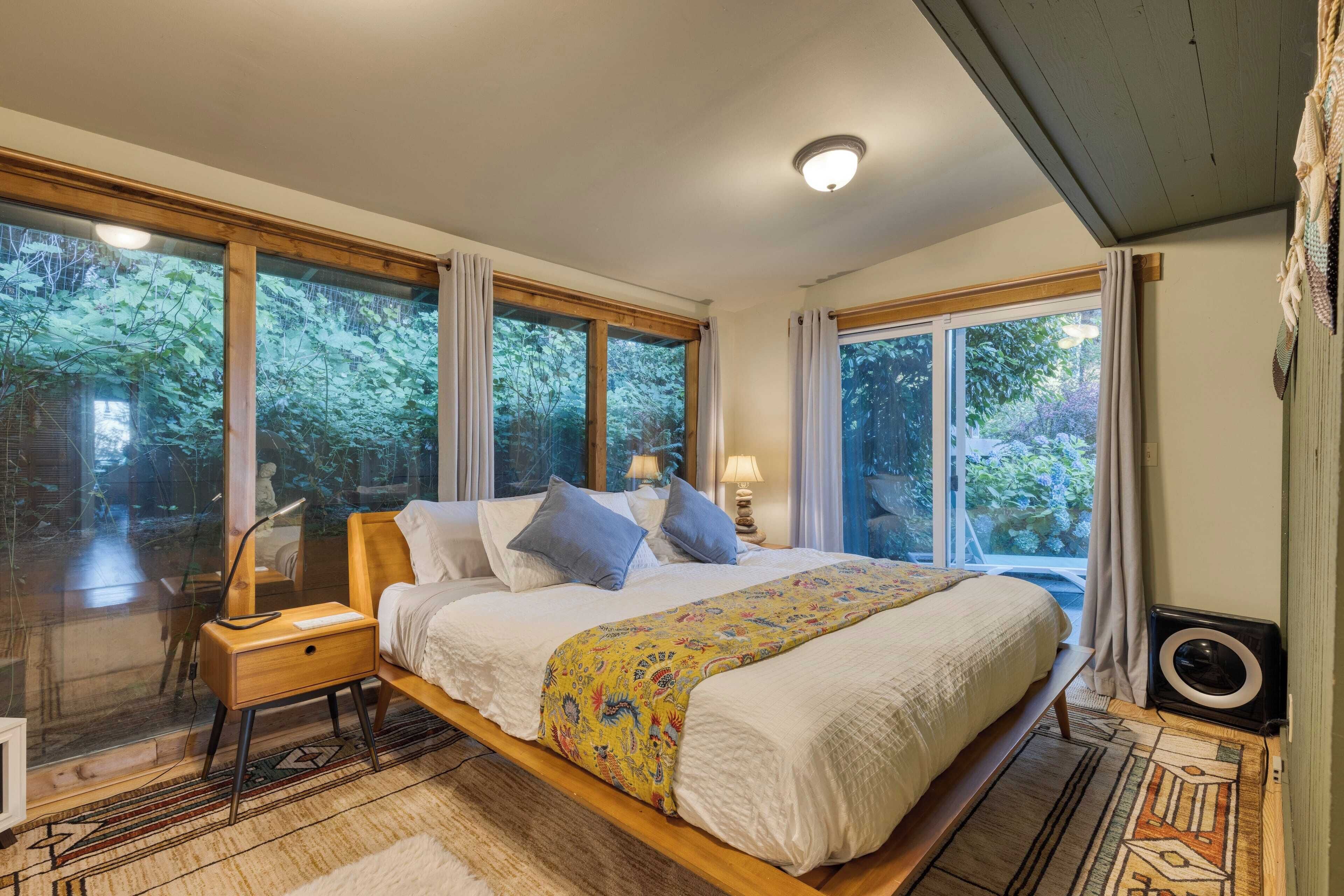 A bedroom with a large bed, wooden headboard, and a view of trees through large windows.