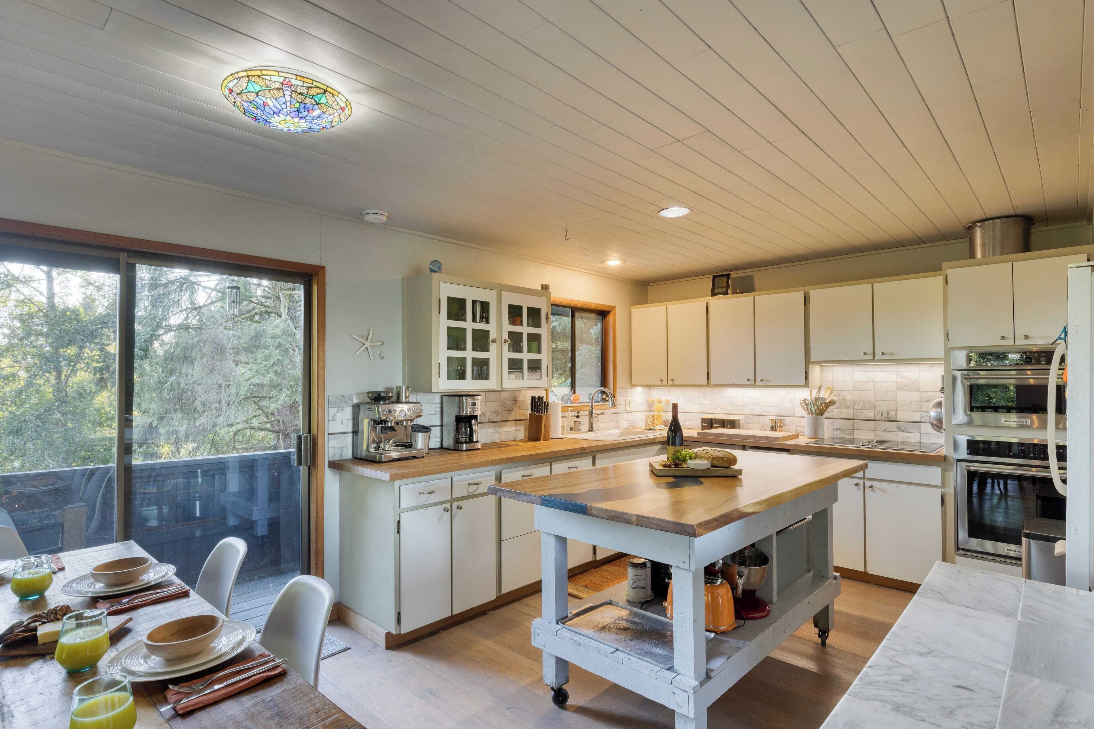 A modern kitchen with white cabinets, a large island, and a view through the window.