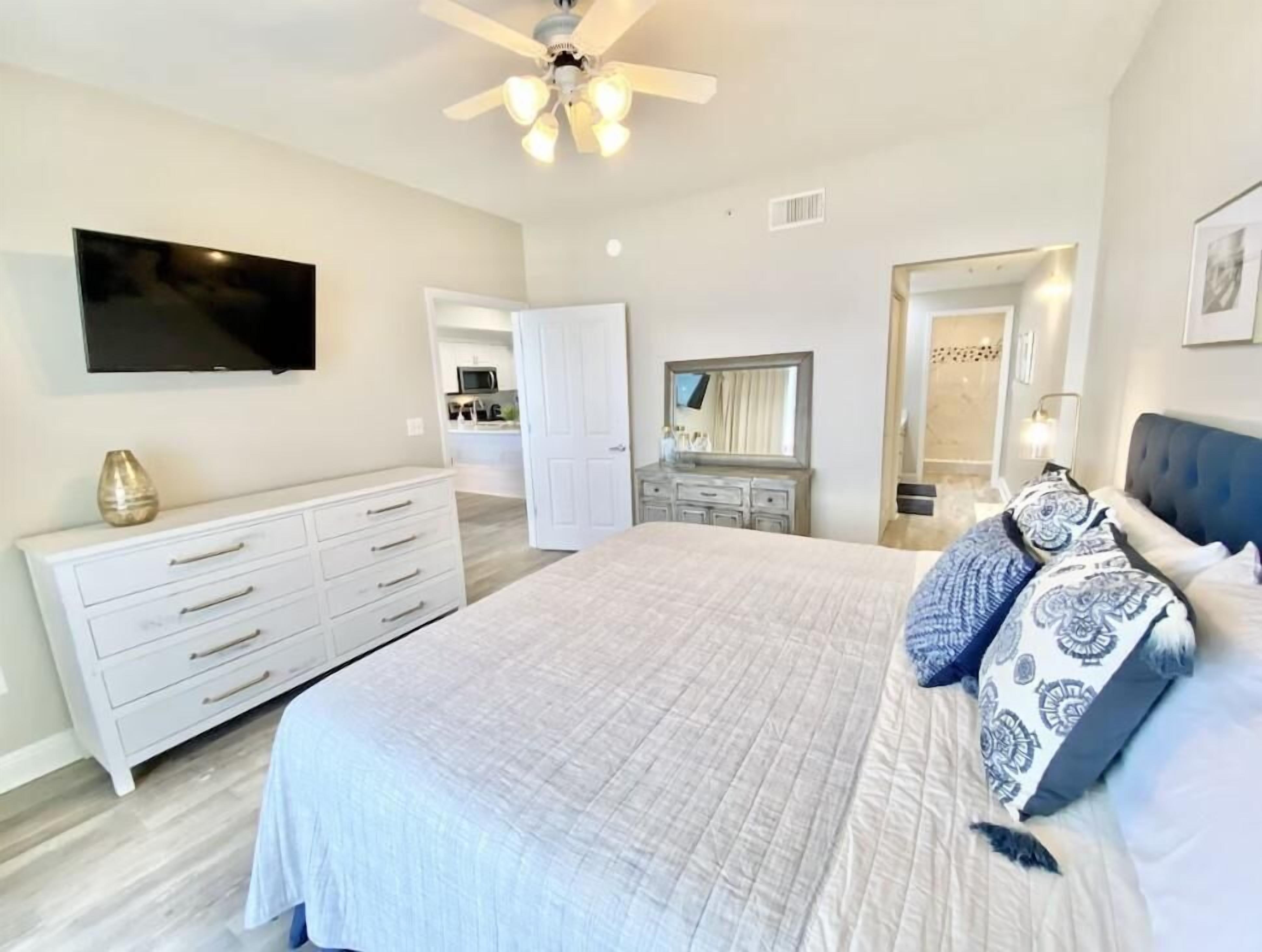 Une chambre moderne avec un grand lit, une commode, un téléviseur, un ventilateur de plafond et une salle de bain visible à travers une porte ouverte.