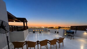 Una terraza en la azotea con vista a la ciudad al atardecer, que incluye un ventilador, sillas y una parrilla.