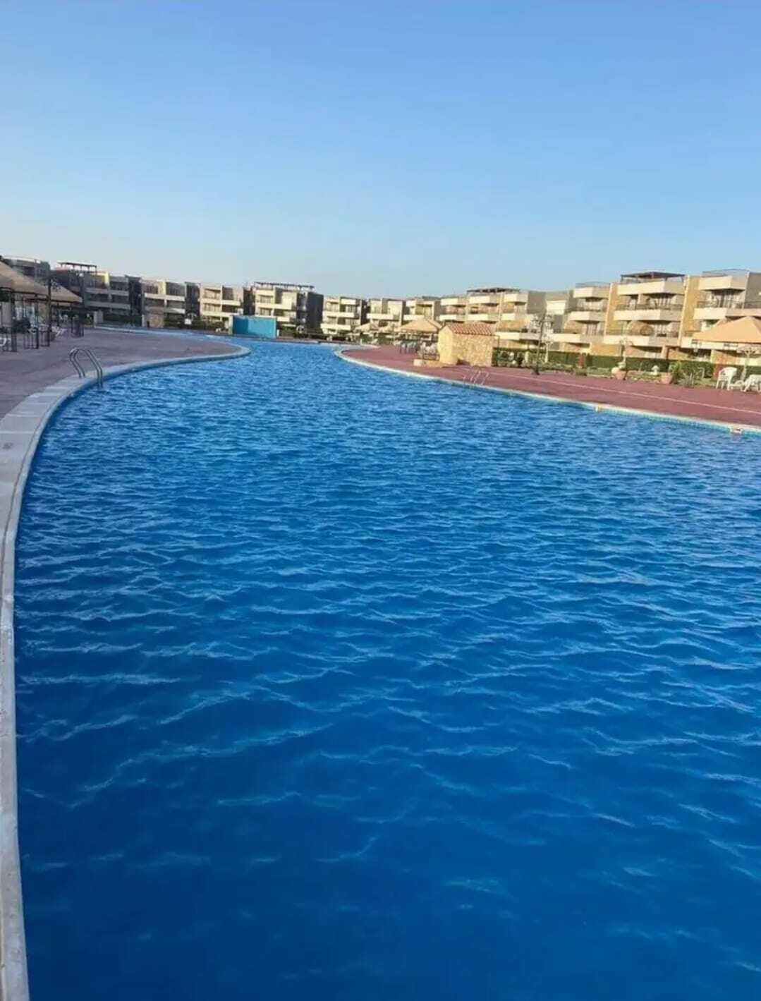 A swimming pool with a curved edge, surrounded by a paved walkway and a row of residential buildings.