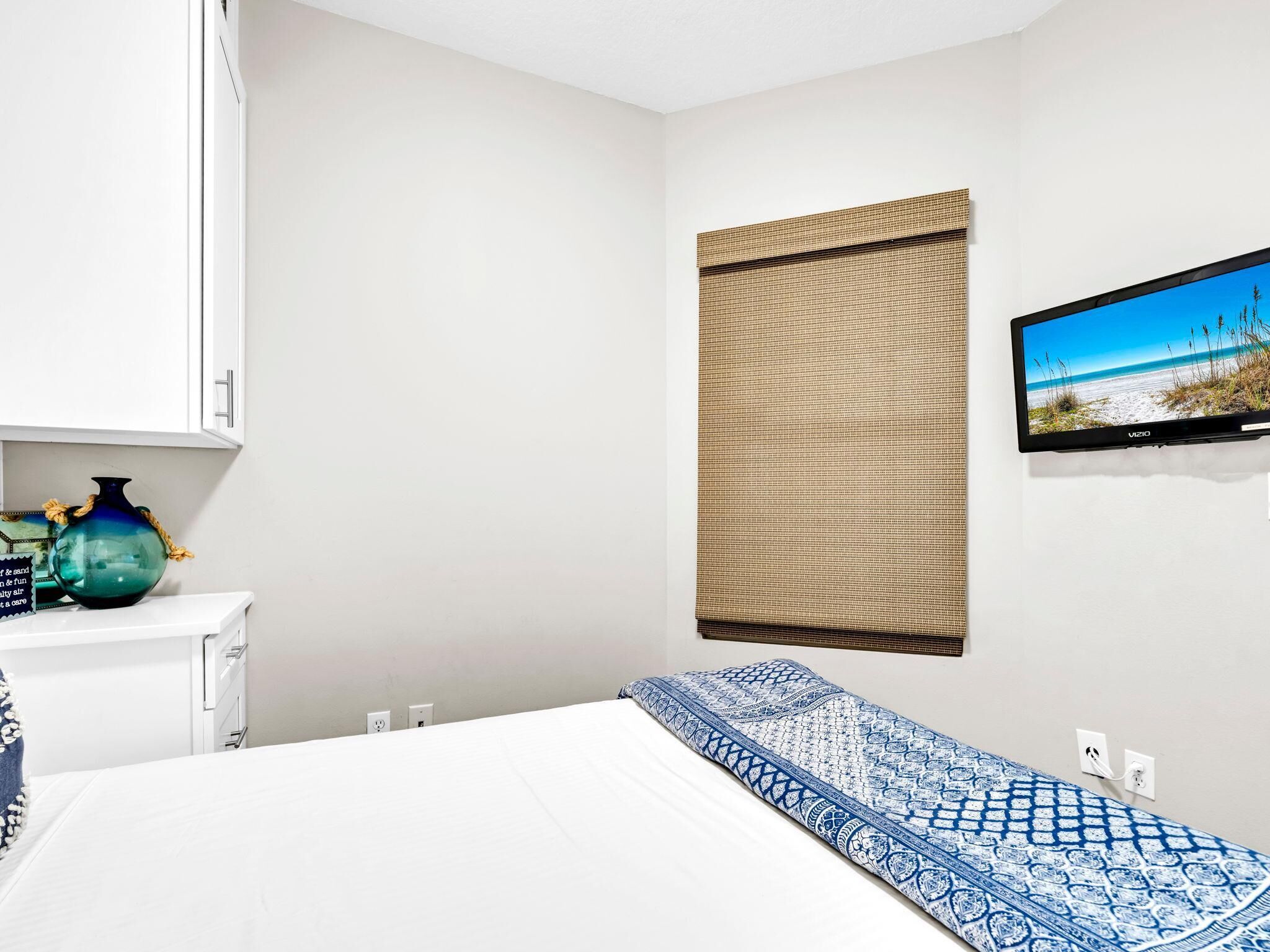 A hotel room with a bed, a television mounted on the wall, a window with a view of the beach, and a small table with decorative items.