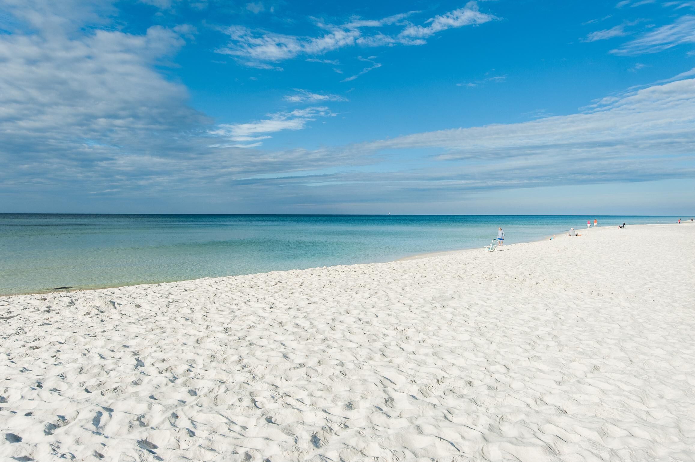 Une vaste plage de sable blanc, bordée d’une eau d’un bleu limpide, et où l’on aperçoit quelques personnes au loin.