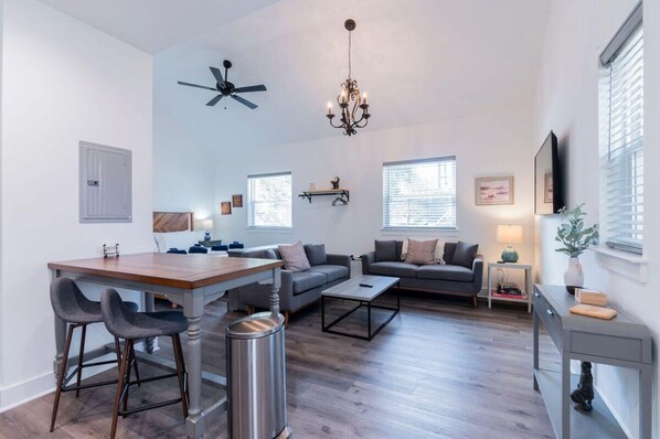 A modern living room with a dining area, grey sofas, a chandelier, and a ceiling fan. - Charming Old Town Loft, Nestled in the Heart of Historic Bluffton, SC (Bluffton)