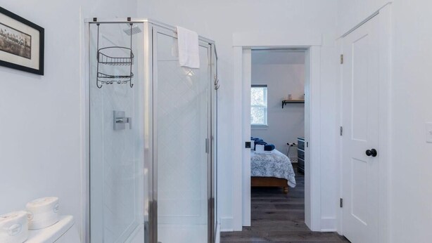 A hallway leading to a bedroom with a bed, a glass shower enclosure, and a white door. - Charming Old Town Loft, Nestled in the Heart of Historic Bluffton, SC (Bluffton)