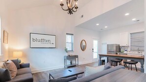 A modern living room with a sofa set, a coffee table, and a dining table. There is a kitchen area with a refrigerator and a sink. A chandelier hangs from the ceiling, and a framed sign on the wall reads "bluffton traveler." - Charming Old Town Loft, Nestled in the Heart of Historic Bluffton, SC (Bluffton)