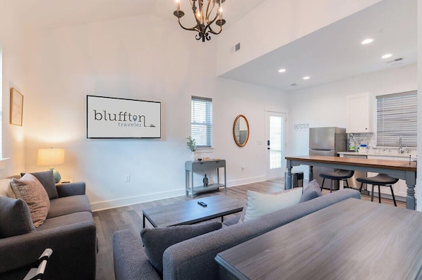 A modern living room with a sofa set, a coffee table, and a dining table. There is a kitchen area with a refrigerator and a sink. A chandelier hangs from the ceiling, and a framed sign on the wall reads "bluffton traveler." - Charming Old Town Loft, Nestled in the Heart of Historic Bluffton, SC (Bluffton)